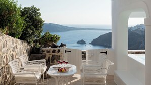 A terrace with white wicker furniture, a table with fruits and glasses, and a view of the sea and hills.