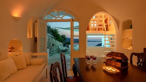 A cozy room with a stone wall, a white sofa, a dining table with chairs, and a view of the sea.