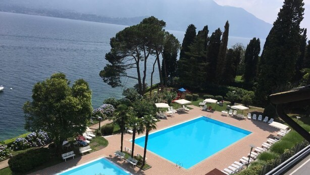 Une piscine entourée d’arbres, avec des chaises longues et des parasols, offrant une vue sur un lac et des montagnes.