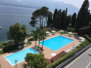 Una piscina con sillones de playa y sombrillas, rodeada de árboles y vista a un lago y montañas.