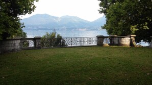 Une zone herbeuse agrémentée d’une clôture en métal décorative et de piliers en pierre, d’où l’on peut admirer un plan d’eau et des montagnes lointaines.