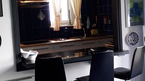 Une pièce moderne avec un grand miroir, deux chaises noires, un sol carrelé bleu et une vue sur un coin cuisine avec une fenêtre et des rideaux.