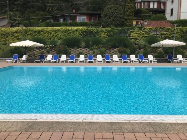 A swimming pool with lounge chairs and umbrellas, surrounded by greenery and buildings.