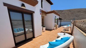 Eine sonnendurchflutete Terrassenanlage im Freien mit weißen Loungesesseln und einer weißen Badewanne, umgeben von einem weißen Gebäude mit Fliesenboden und Blick auf eine gebirgige Landschaft.