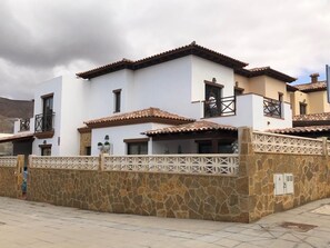A two-story building with a white facade and terracotta roof tiles. It features a stone wall with a decorative lattice design and a small balcony with a metal railing. - ABSOLUTE TRANQUILITY, COMFORTABLE AND CENTRAL LOCATION. (Tuineje)