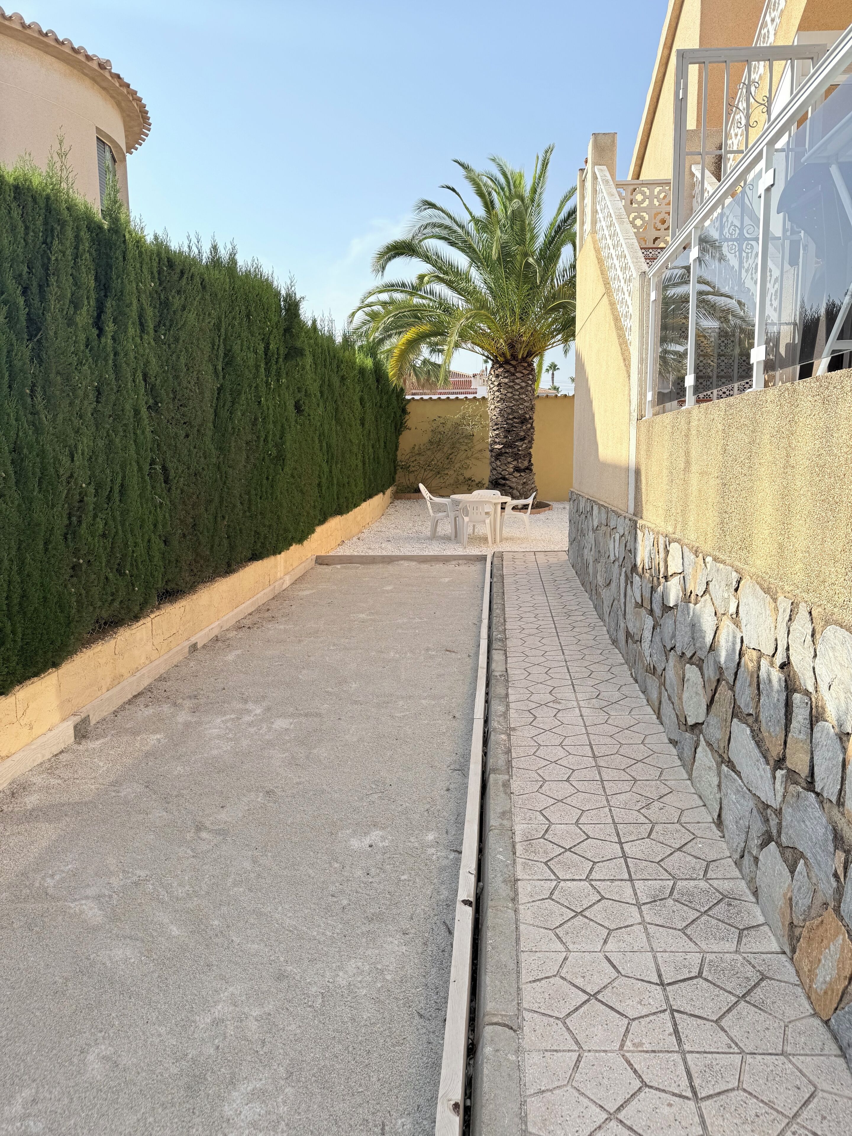 A paved pathway flanked by a stone wall and a hedge, leading to a building with a tiled entrance.
