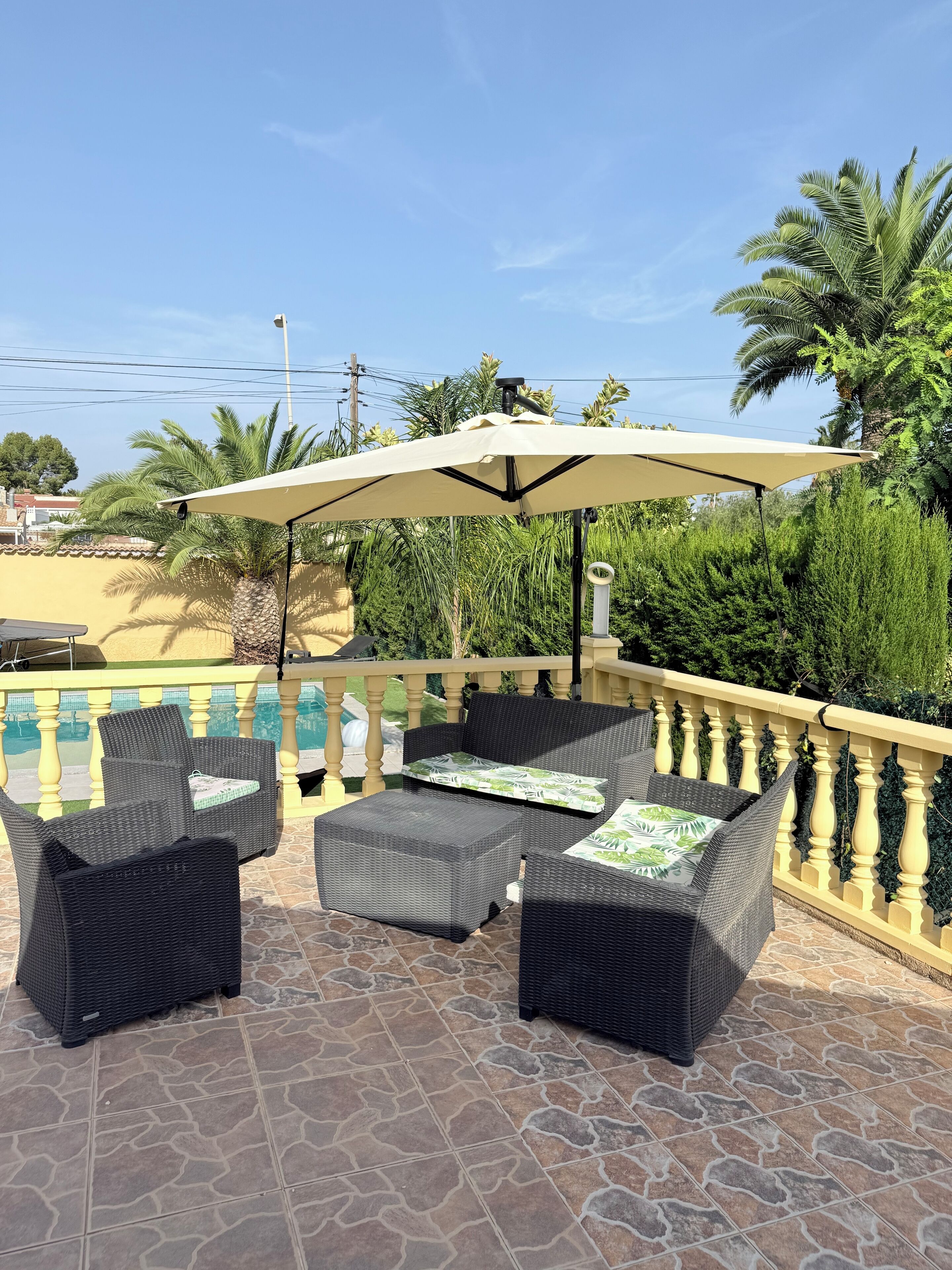 Pátio externo com móveis de vime e um guarda-sol grande, com vista para uma piscina e vegetação exuberante.