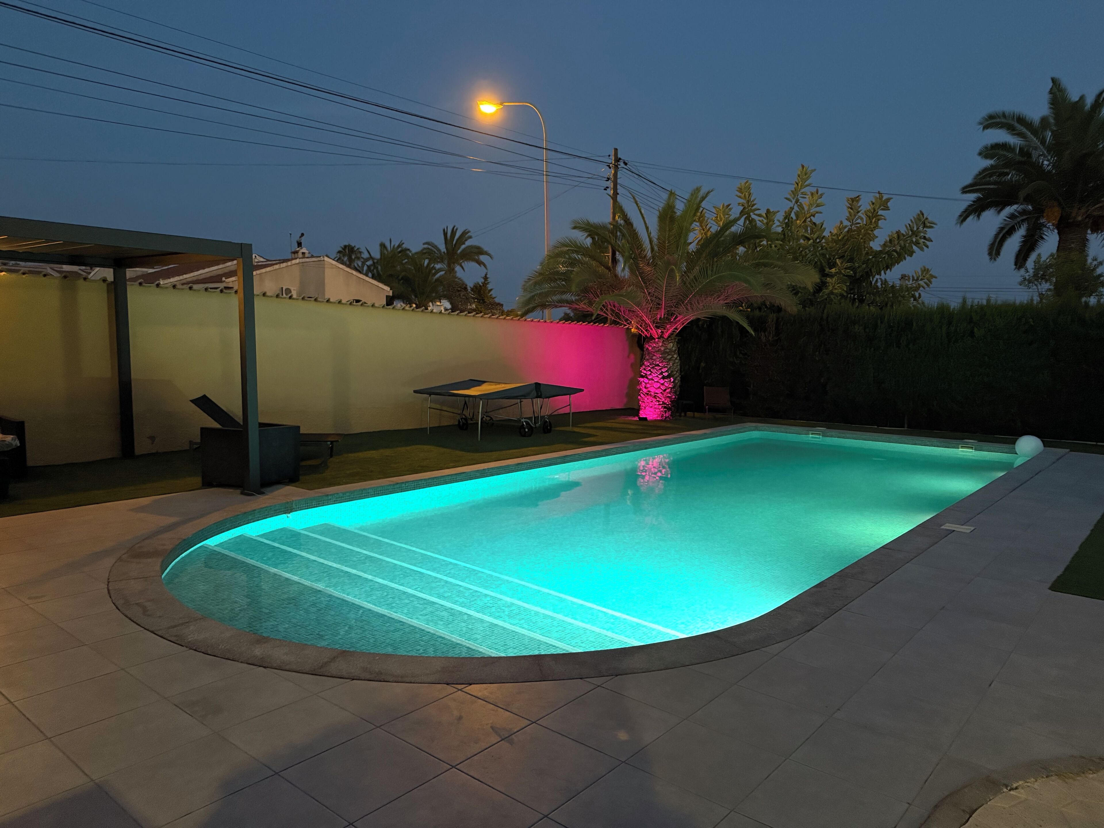 A swimming pool with a tiled deck, a covered seating area, and a street lamp.
