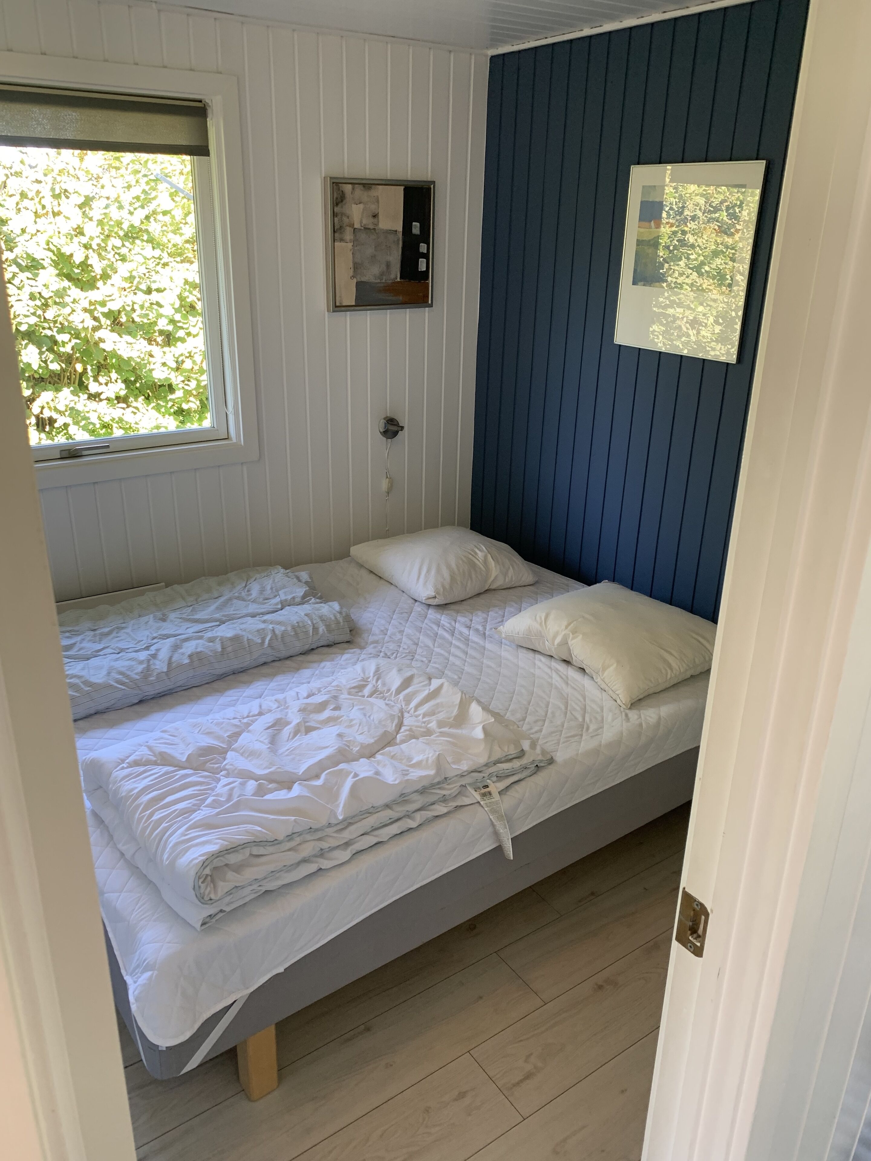 Een eenpersoonsbed met wit beddengoed in een kamer met houten vloeren, een raam met zonwering en een blauwe muur met een ingelijst beeld.