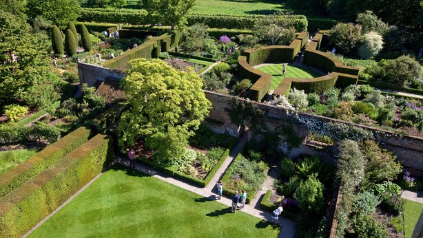 Une vue aérienne d’un jardin magnifiquement entretenu, avec des haies géométriques, des allées et une grande variété d’arbres et d’arbustes.