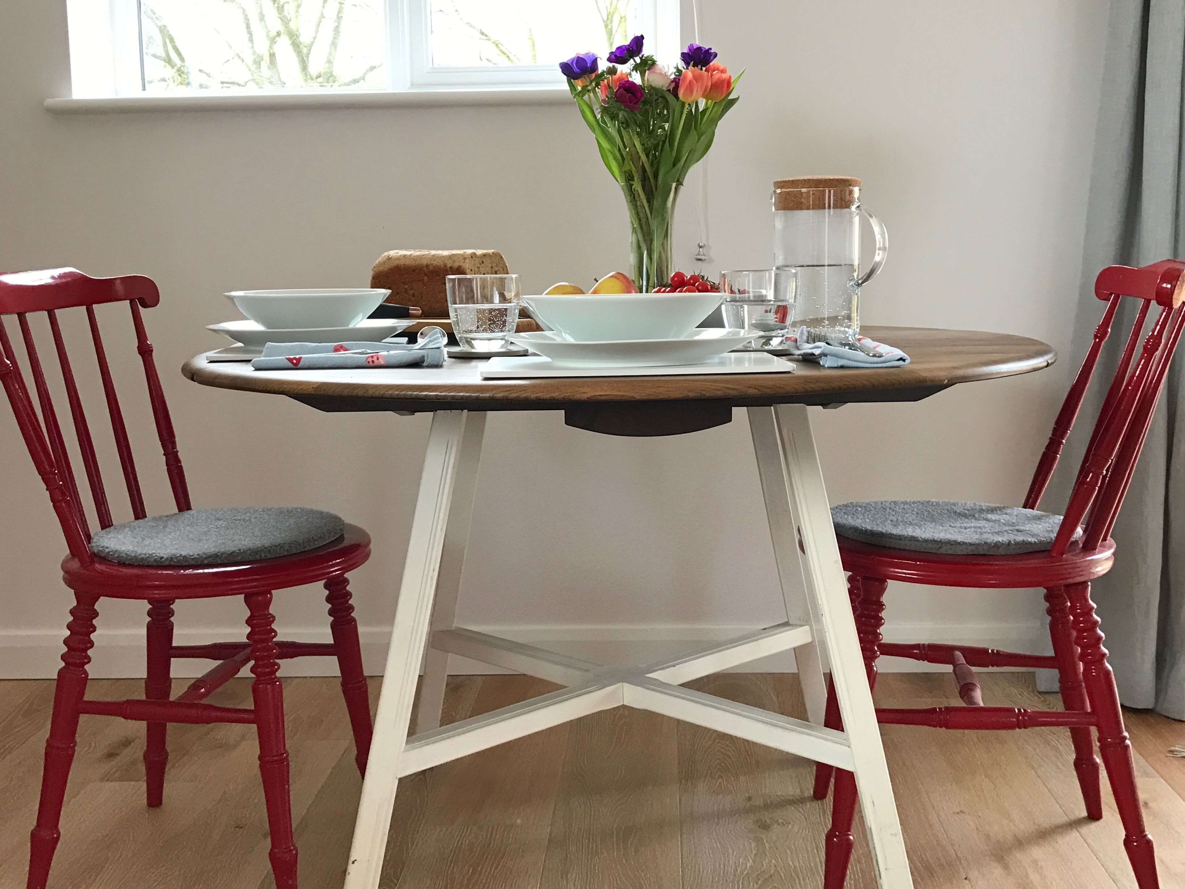 A dining table with chairs, a vase of flowers, plates, a pitcher, and a loaf of bread.