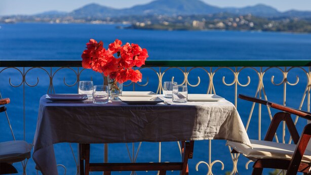 A table with a tablecloth, a vase with red flowers, and glasses on a balcony overlooking a body of water and distant mountains. - Vill Pergola Elli, holiday apartment in Corfu, Greece with stunning sea view! (Corfu)