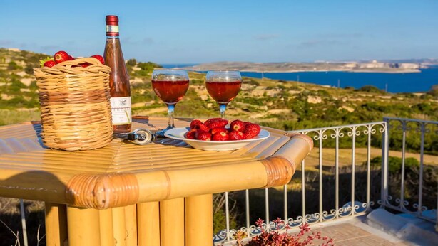 A table with a bottle, glasses, and strawberries, overlooking a coastal landscape. - Great Location: FREE Gozo transfers, WIFI, Bicycles & BBQ  – NO Car required! (Ghajnsielem)