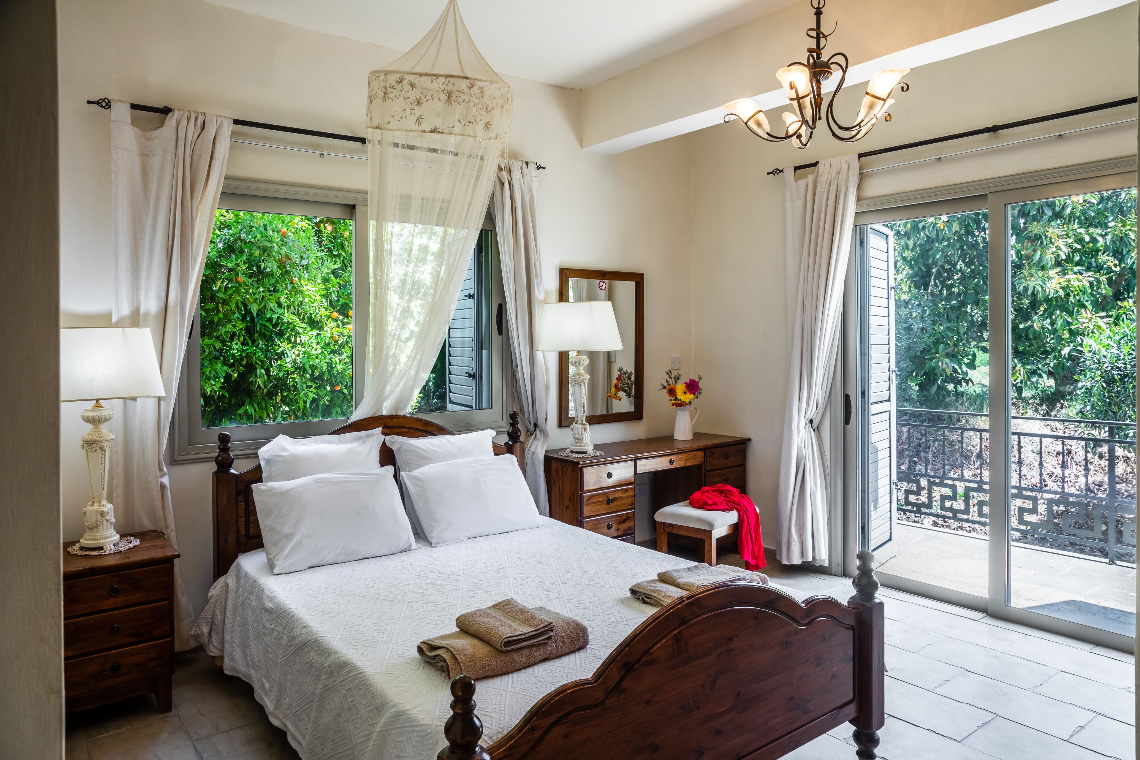 A bedroom with a large bed, a nightstand, a dresser, a chair, and a balcony with a view of greenery.