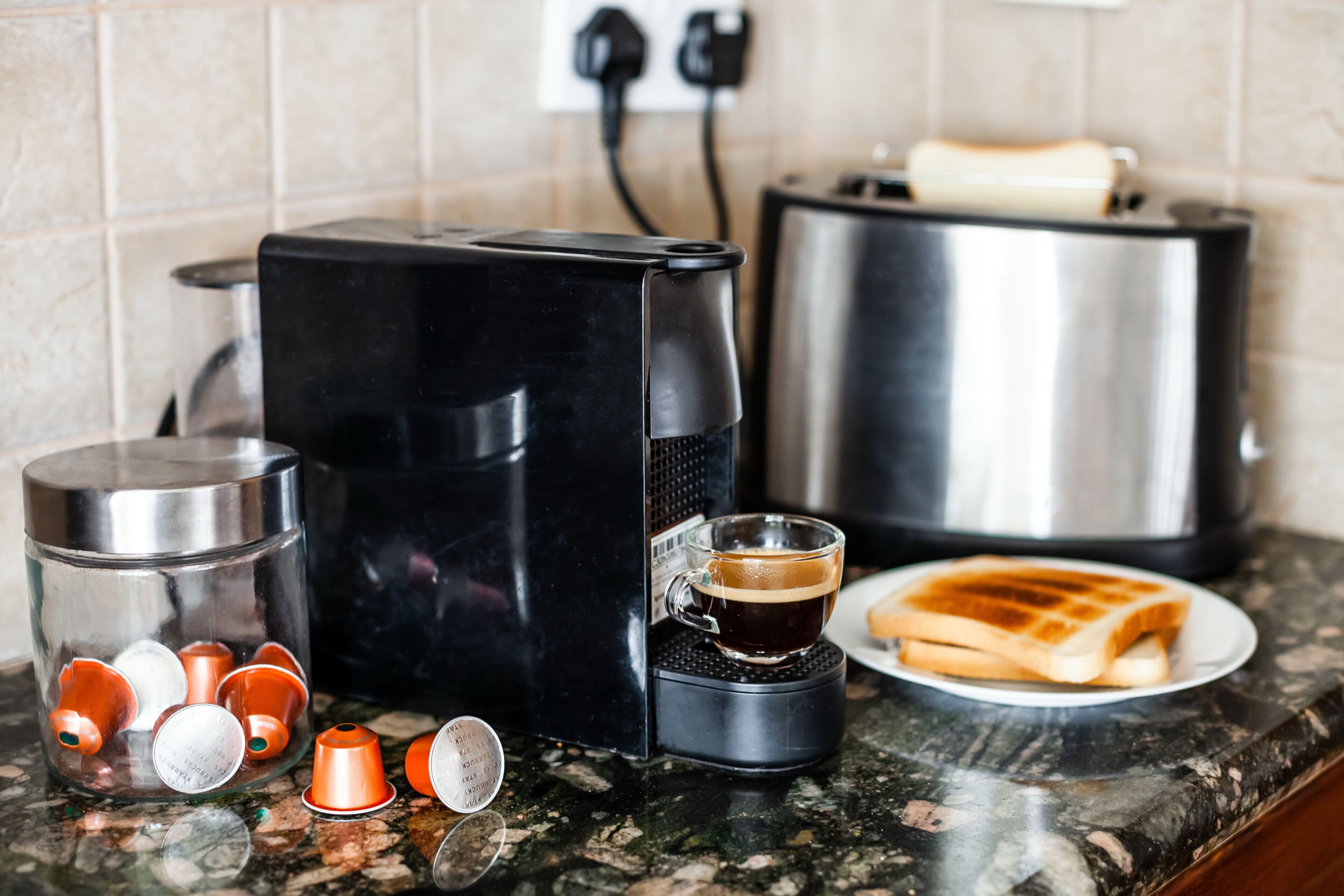 Ett kök med en kaffebryggare, en brödrost, en glasbehållare med kaffekapslar och en tallrik med rostat bröd.