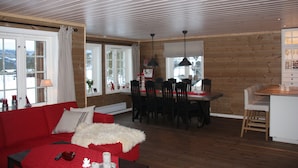 A cozy living room with a red sofa, a white furry throw, and a dining area with wooden walls and a kitchen counter. - Luxury cottage with panoramic views. Proximity to summer and winter activities. (Hemsedal)