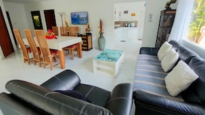 A modern living room with a black leather sofa, wooden dining chairs, a glass coffee table, and a TV mounted on the wall. - ⭐⭐⭐⭐⭐Villa 4 bedrooms with private pool 5 minutes beaches and Walking Street. (pattaya)
