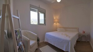Ein Schlafzimmer mit einem Bett, einem Sessel, einem Nachttisch, einer Lampe, einem Fenster und einem an der Wand befestigten Regal.
