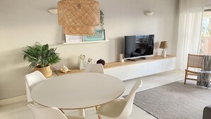 A modern living room with a round dining table, white chairs, a flat-screen TV, a woven pendant light, and a bookshelf with decorative items. - Saltwater Apartment - Sea View (Portimão)