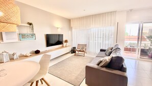 A modern living room with a flat-screen TV, a sofa, a dining table, and a large window with sheer curtains. - Saltwater Apartment - Sea View (Portimão)