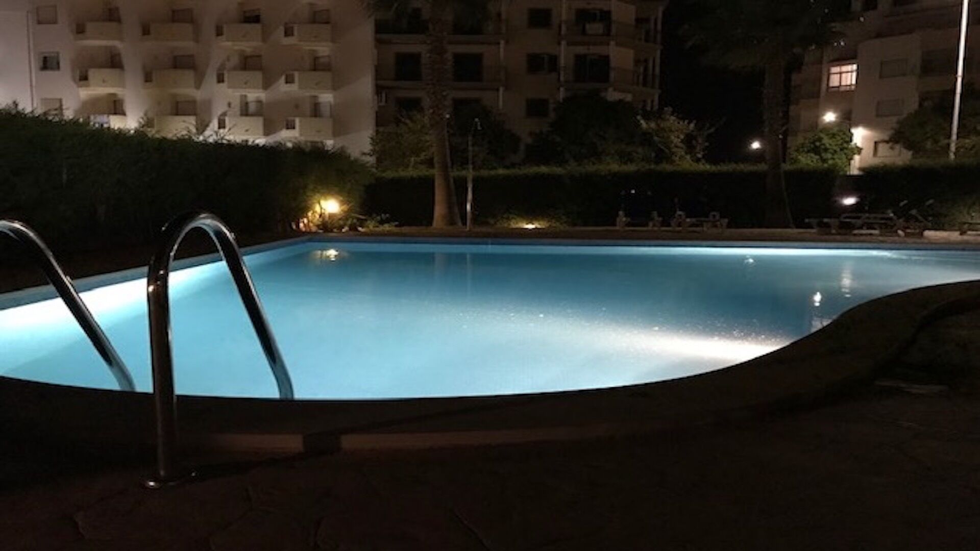 En swimmingpool om natten med en metalstige, omgivet af en hæk og bygninger i baggrunden.