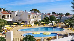 Ein Resort mit einem großen Schwimmbecken, umgeben von Liegestühlen und Sonnenschirmen, weiße Gebäude mit roten Dächern und einem strahlend blauen Himmel.