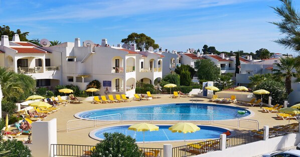 A resort with a large swimming pool surrounded by sun loungers and umbrellas, white buildings with red roofs, and a clear blue sky.