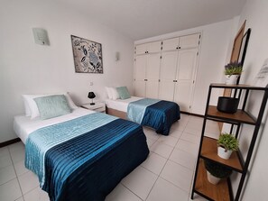A bedroom with two beds, a wardrobe, a shelf with plants, and a wall-mounted light.