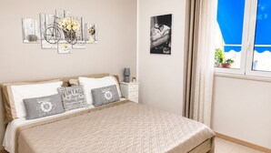 Ein Schlafzimmer mit Bett, Kissen, Nachttisch, Lampe, einem gerahmten Bild und einem Fenster mit Vorhängen.