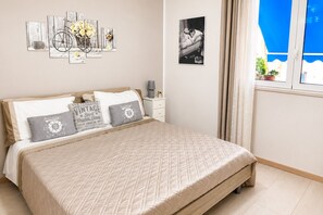 Et soveværelse med en seng, puder, et natbord, en lampe, et billede i ramme og et vindue med gardiner.