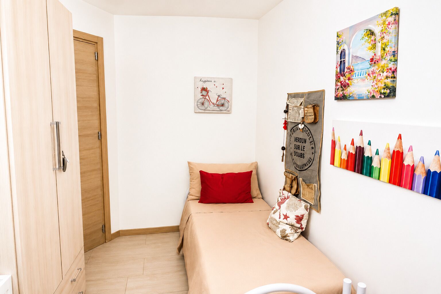 Un dormitorio pequeño con una cama, un armario de madera, una puerta, una pared con un cuadro enmarcado y una pared con lápices de colores.