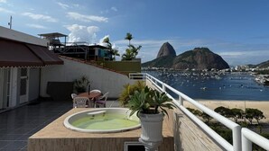 Terraza en la azotea con jacuzzi, zona de estar y vista a una bahía con barcos y una montaña.