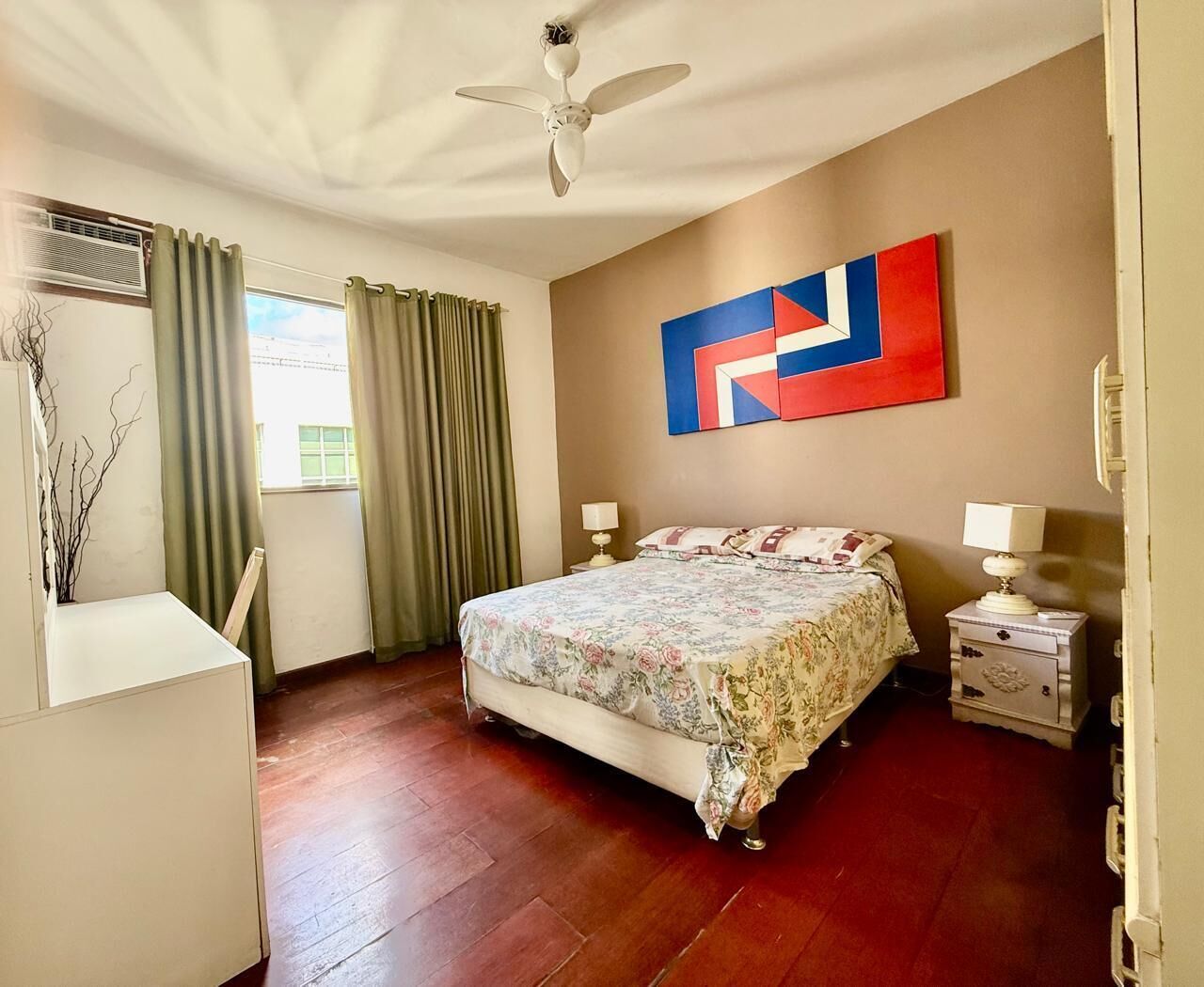 A bedroom with a bed, two bedside tables, a ceiling fan, a window with curtains, and a wall-mounted artwork.