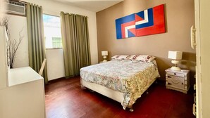 Un dormitorio con cama, dos mesitas de noche, ventilador de techo, ventana con cortinas y una obra de arte colgada en la pared.