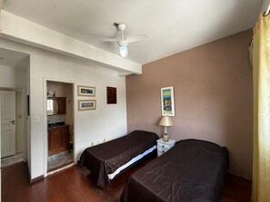 A room with two single beds, a ceiling fan, a small table with a lamp, and a door leading to a kitchen area.