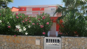 Un mur de pierre avec un portail en métal, un bâtiment rouge et blanc avec un toit en tuiles, et des plantes en fleurs.