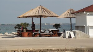 Un café situé en bord de mer, avec des tables en bois et des parasols en chaume, offrant une vue sur l’océan.
