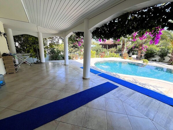 A covered outdoor area with a swimming pool, surrounded by plants and a pergola.