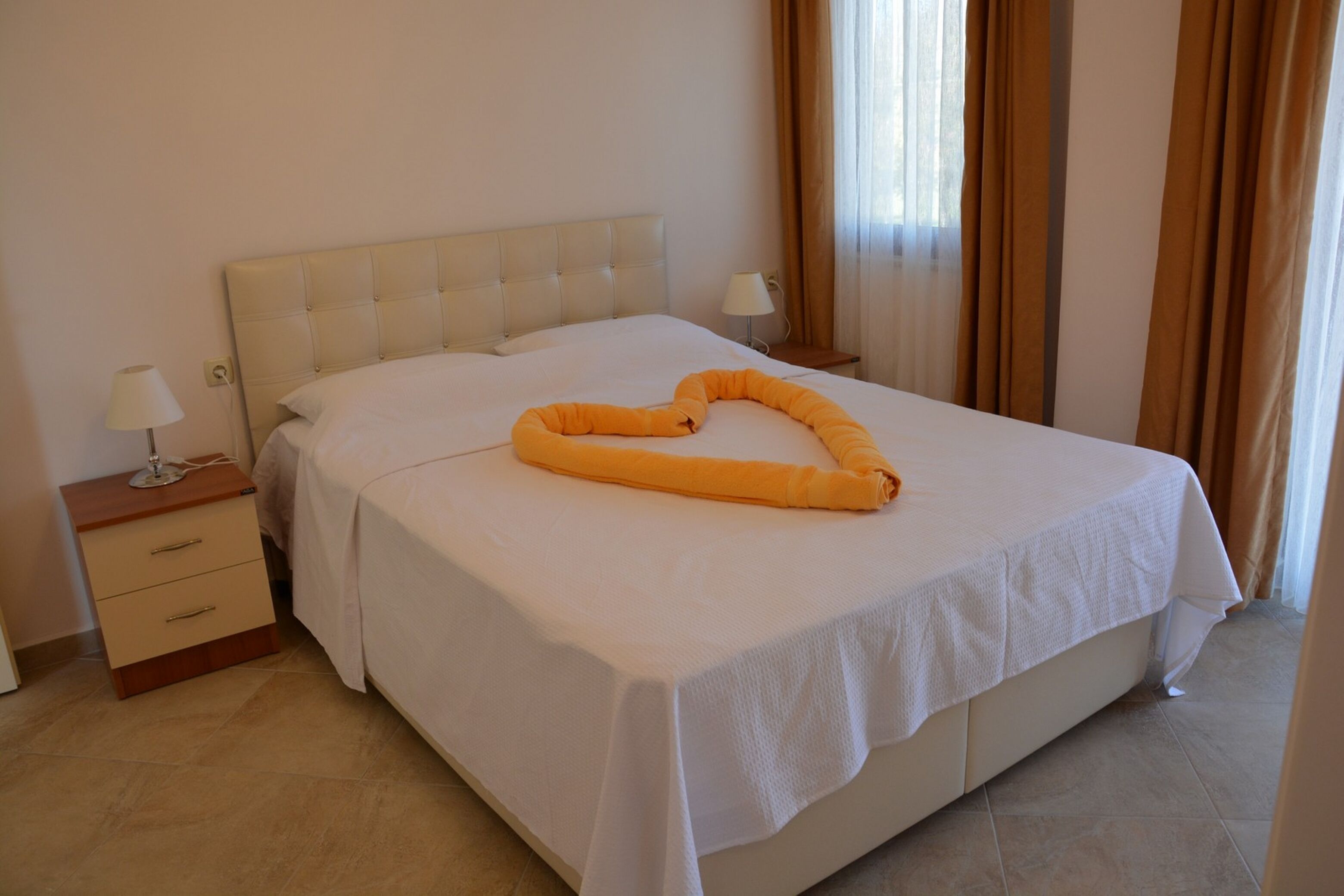 Un letto rifatto con cura, un asciugamano a forma di cuore al centro, un comodino con una lampada e una finestra con tende.