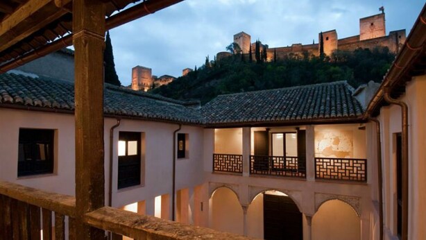 Un patio con barandilla de madera, arcos y una vista de un castillo al fondo.