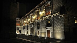 Un edificio histórico con detalles arquitectónicos ornamentados, incluyendo un balcón y numerosas ventanas, iluminado por la noche.