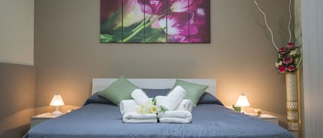 Una cama bien tendida con una colcha azul, dos almohadas verdes y un arreglo floral en la mesita de noche.