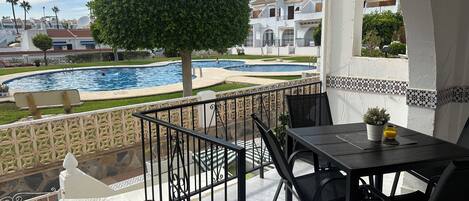 Un balcón con vista a una piscina, un árbol y un edificio con paredes blancas y detalles azules.