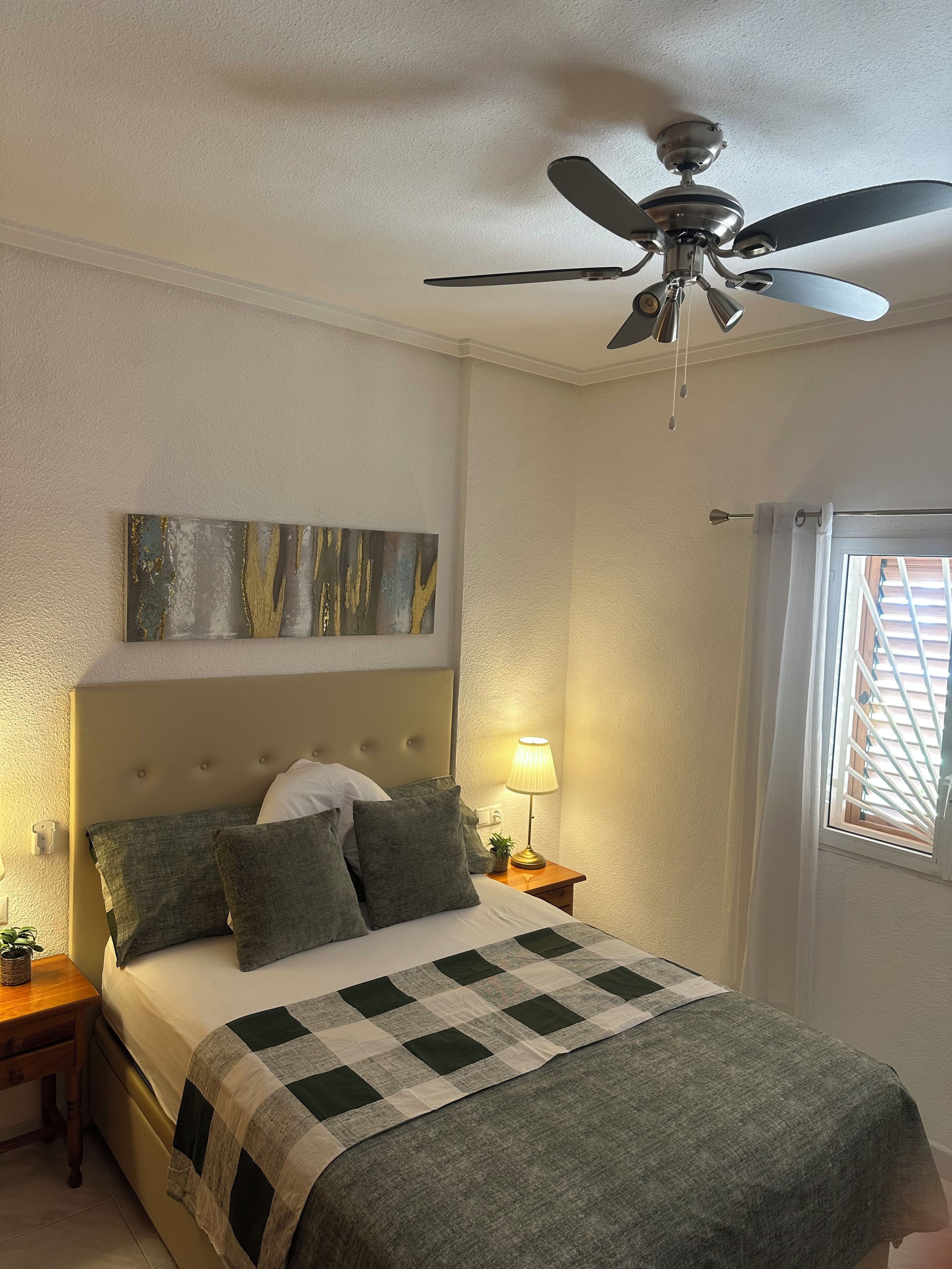A bedroom with a bed, a ceiling fan, a nightstand with a lamp, and a window with blinds.
