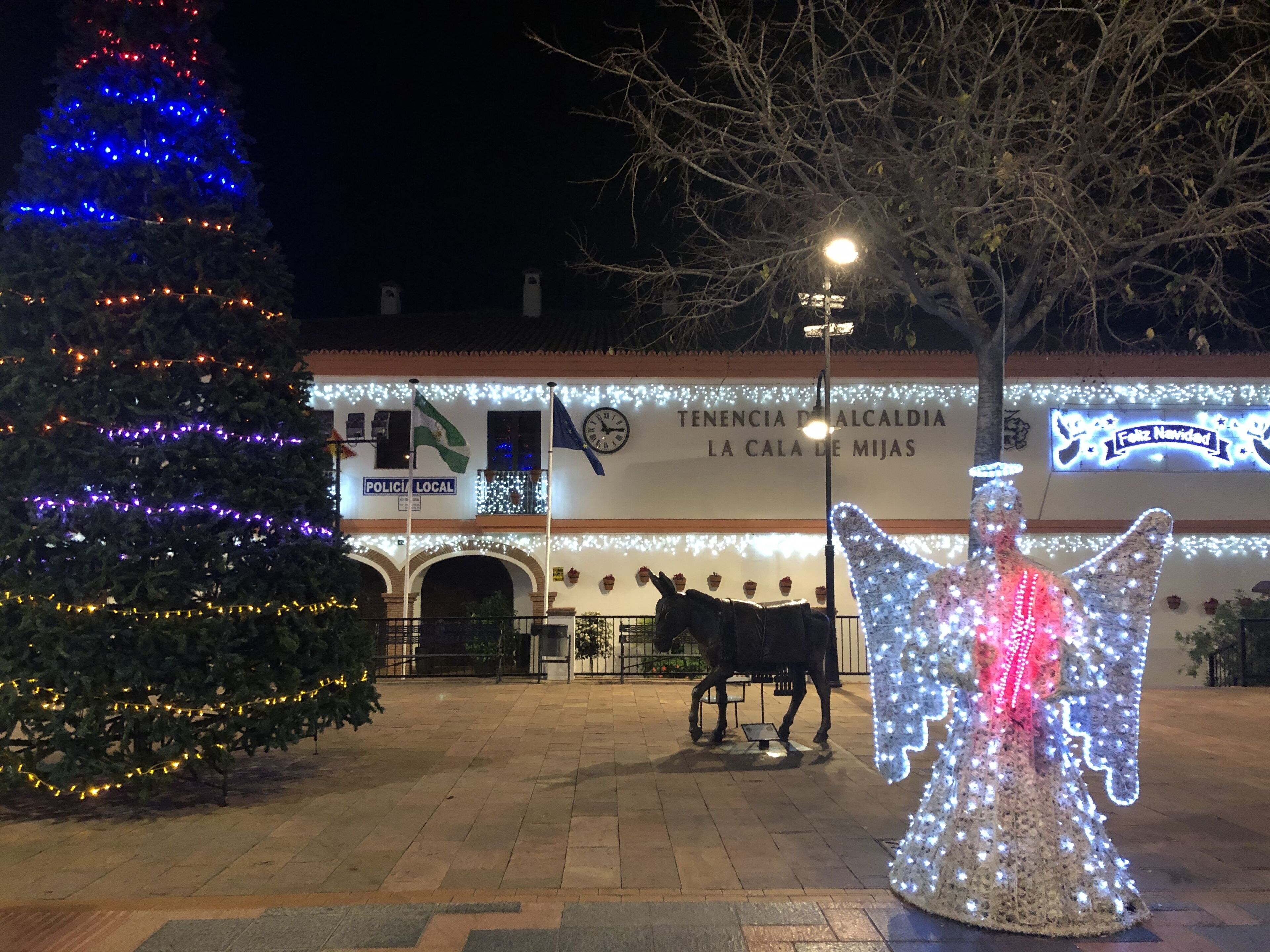 Una plaza con un árbol de Navidad, un carruaje tirado por caballos y una estatua iluminada de un ángel.