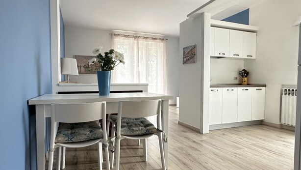 A dining area with a white table and chairs, a vase with flowers, and a kitchen with white cabinets. - The Seagull apartment Sirmione | Spa & Garda Lake (Sirmione)
