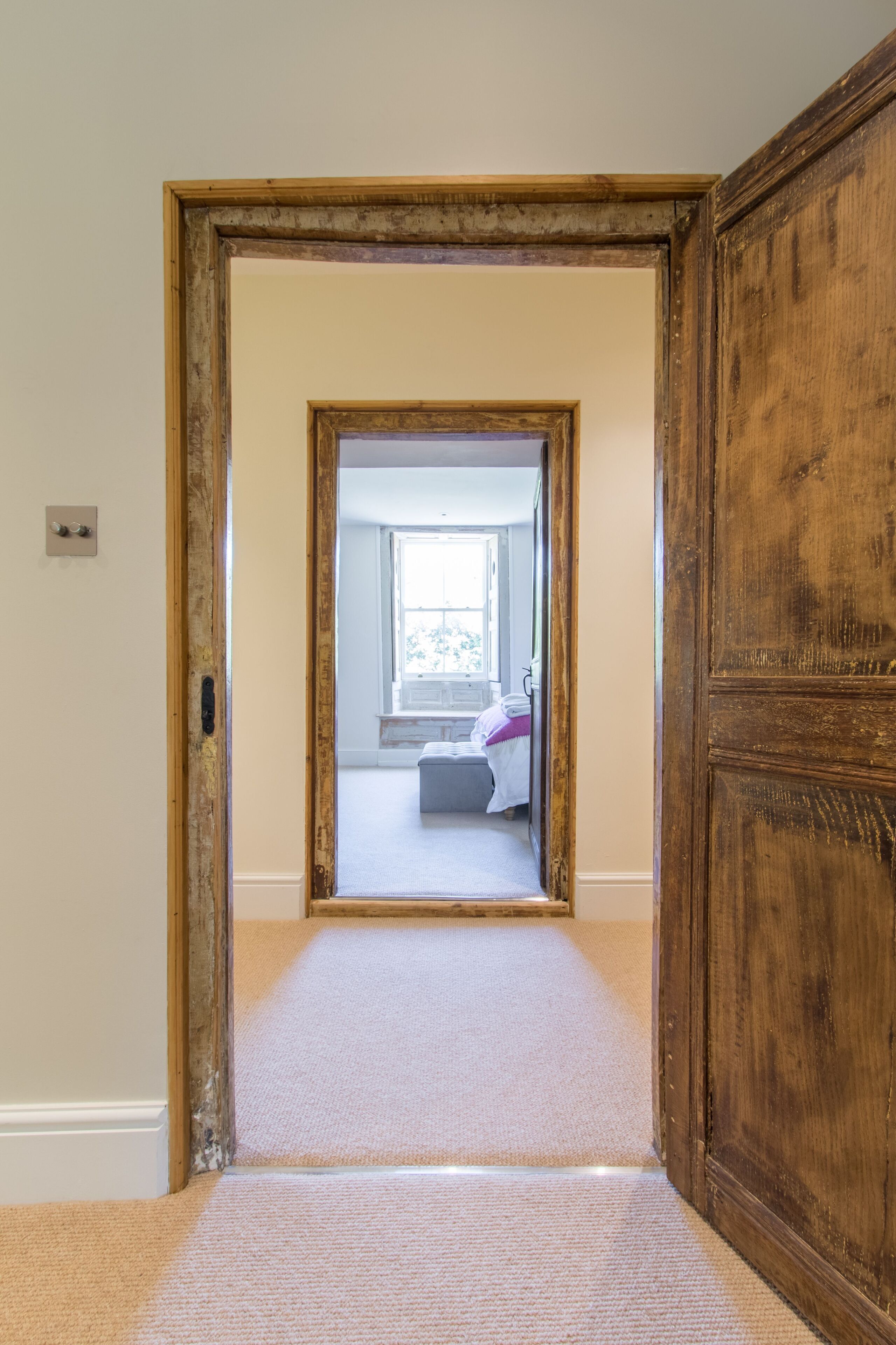 A hallway leading to a bedroom with a large window, carpeted floor, and wooden door frames.