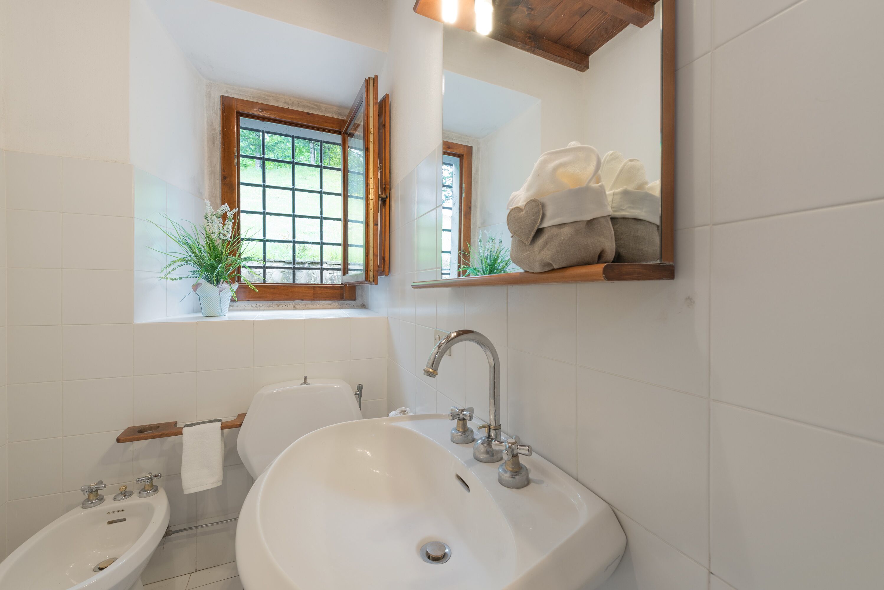 A bathroom with a white sink, a bidet, a window with a view of greenery, and a towel rack with towels.