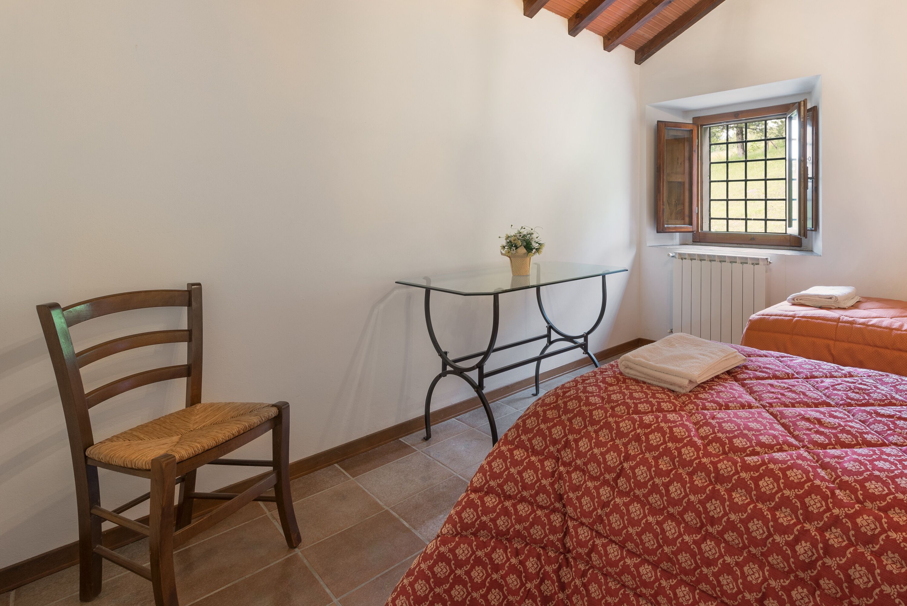 A bedroom with a wooden chair, a small table with a plant, a window with a view, and a bed with a red patterned bedspread.