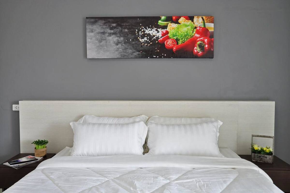 Ein ordentlich bezogenes Bett mit weißen Leinen, ein grauer Kopfteil und ein an der Wand hängendes Bild von roten Paprika und anderem Gemüse.